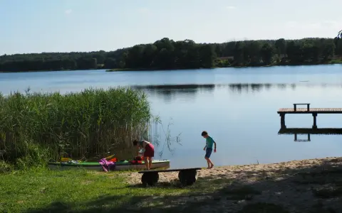 Dzieci szykują kajak nad spokojnym jeziorem w gminie Morąg. W tle las i drewniane molo.