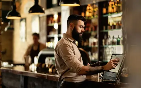 Hombre con barba gestionando un bar, usando una tablet y una pantalla táctil para procesar pagos.