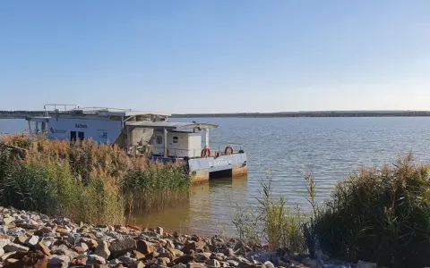 Ein Katamaran namens "Barbara" liegt am Ufer des Schlabendorfer Sees, umgeben von Schilf und Steinen.