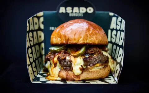 Saftiger Burger mit geschmolzenem Käse und Gurken, präsentiert in einer "ASADO BURGERS" Box. Der beste Burger Kölns wartet!