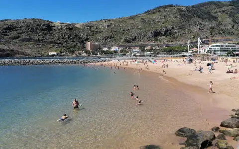 Sonniger Strand in Machico, Madeira, mit vielen Menschen, die im klaren Wasser planschen und sich am Sandstrand sonnen.