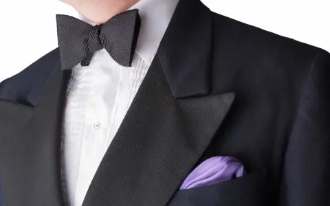 A man wears a black tuxedo with a pleated white shirt and a black bow tie. A purple pocket square adds a pop of color. This outfit answers the question: can you wear a tie with a tux? (No, a bow tie is traditional.)