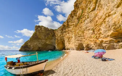 Ein Boot liegt am Korfu Strand vor einer beeindruckenden Felswand. Türkises Wasser und ein sonniger Himmel laden zum Verweilen ein.