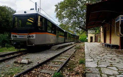 Ein Zug nach Griechenland steht auf den Gleisen neben einem alten Bahnhofsgebäude. Herbstlaub bedeckt den Bahnsteig.
