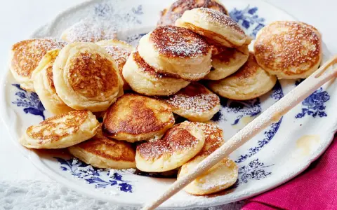 Ein Teller voller goldbrauner Poffertjes, bestäubt mit Puderzucker. Dieses Poffertjes Rezept ist ein Genuss!