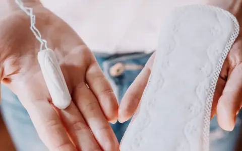 Dłonie trzymają tampon i podpaskę. Czasem pojawia się plamienie zamiast okresu, wtedy wybór jest ważny.
