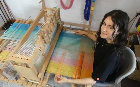 Une jeune femme souriante, passionnée par son tissier métier, travaille sur un métier à tisser en bois. Les fils colorés créent une œuvre vibrante.