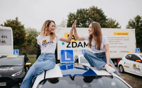 Dwie uśmiechnięte dziewczyny świętują sukces na dachu samochodu szkoleniowego. W tle ciężarówka z informacją o kursach na różne kategorie prawa jazdy.