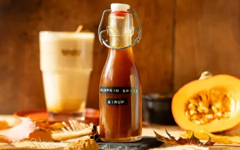 Flasche mit goldbraunem Pumpkin Spice Sirup, daneben ein Latte Macchiato, Kürbishälften und Herbstlaub.