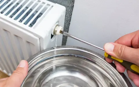Osoba odkręca odpowietrznik grzejnika śrubokrętem, a woda kapie do metalowej miski. To pokazuje, jak odpowietrzyć grzejnik.