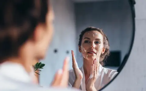 Une femme se regarde dans un miroir, appliquant de la mousse sur son visage pour un double nettoyage.