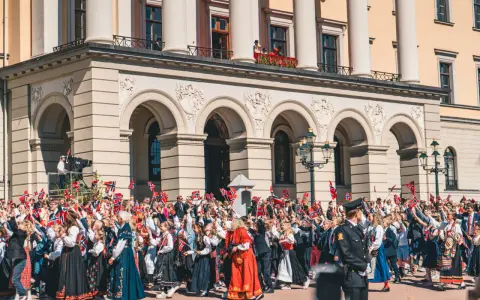 Tłum ludzi, w tym dzieci, świętuje, machając norweskimi flagami. Wiele osób nosi tradycyjny strój norweski, ozdobiony haftami i kolorowymi wzorami.