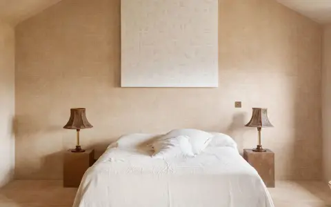 Camera da letto aesthetic con pareti color sabbia, letto bianco e due lampade in vimini. Un quadro astratto completa l'arredo.