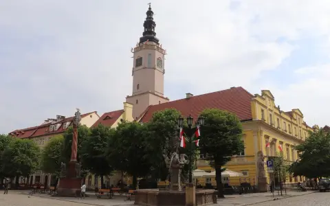 Wieża ratuszowa w Świdnicy góruje nad rynkiem, ozdobiona polskimi flagami. Obok fontanna i zabytkowe kamienice.