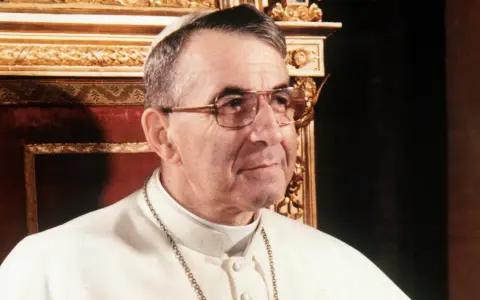 Papież Jan Paweł I, który był papieżem przed Janem Pawłem II, w białej sutannie i okularach.