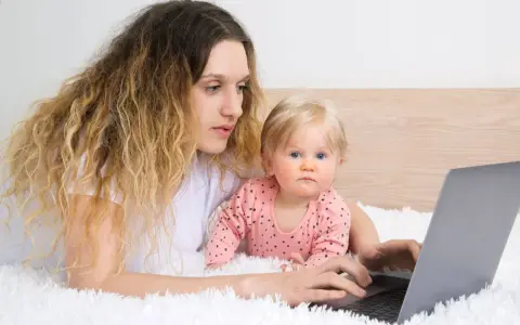 Kobieta z dzieckiem na rękach pracuje na laptopie, zastanawiając się, czy na urlopie wychowawczym można pracować.