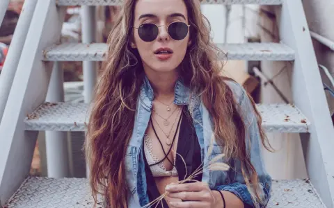 Frau mit langen Haaren und Sonnenbrille sitzt auf einer Metalltreppe. Sie trägt ein offenes Jeanshemd, das sie lässig mit einem Spitzen-Bralette kombiniert.