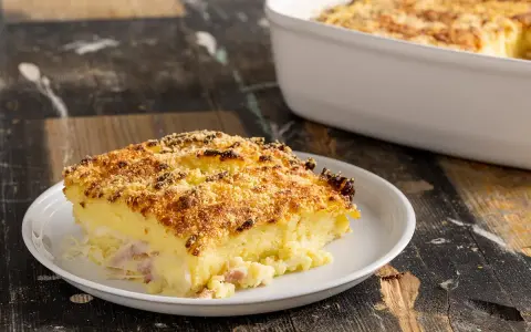 Una fetta di sformato di patate dorato e croccante, con prosciutto e formaggio filante, servita su un piatto bianco.