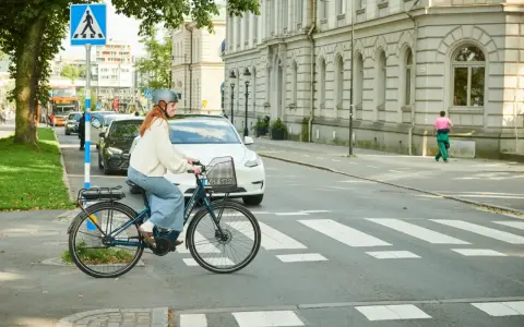 Kvinna på elcykel korsar övergångsställe. Trafik och byggnader i bakgrunden. Elcykel regler är viktiga för säkerheten.