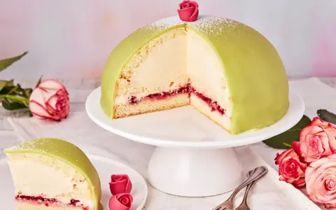 Ein Anschnitt eines grünen Prinzessinnenkuchens mit Sahnefüllung und Himbeermarmelade, verziert mit einer Zuckerrose. Ein Stück liegt auf einem Teller daneben.