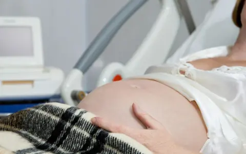 Kobieta w szpitalnym łóżku, z ręką na brzuchu, czeka na poród. Obok monitor KTG, który pomaga ocenić, jak przyspieszyć rozwarcie.