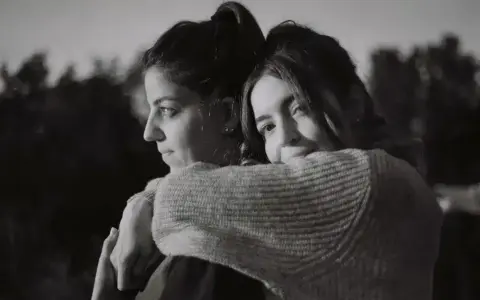 Dos mujeres se abrazan, una mirando al horizonte con **miedo al abandono**, la otra la abraza con ternura.