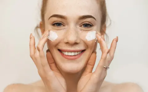 Uśmiechnięta dziewczyna z piegami nakłada krem na twarz. Sudocrem na co? Na suchą skórę, podrażnienia, zaczerwienienia.