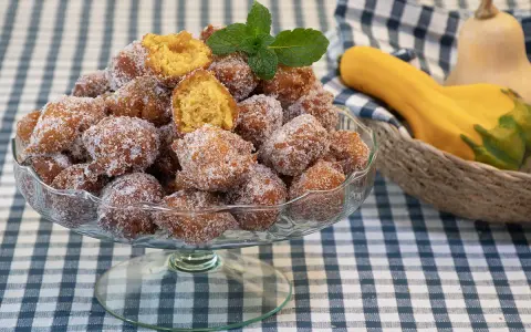 Un tazón lleno de buñuelos de calabaza cubiertos de azúcar, con una ramita de menta encima. Al fondo, calabazas en una cesta.