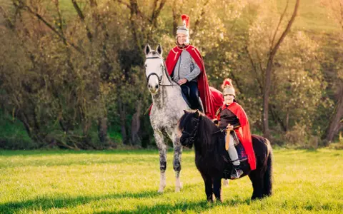 Zwei Reiter in römischer Kleidung, ein Erwachsener auf einem Schimmel und ein Kind auf einem Pony, reiten auf einer sonnigen Wiese. Ein fröhlicher Anblick, wie bei einem Martin Fest.