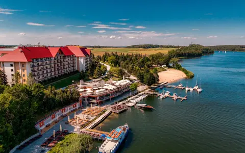 Hotel na wyspie Mikołajki z czerwonym dachem, otoczony zielenią, z mariną pełną łodzi i statkiem wycieczkowym na jeziorze.
