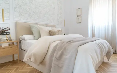 Dormitorios con papel pintado de hojas verdes en el cabecero. Cama con ropa de cama blanca y manta gris. Mesita de noche de madera.