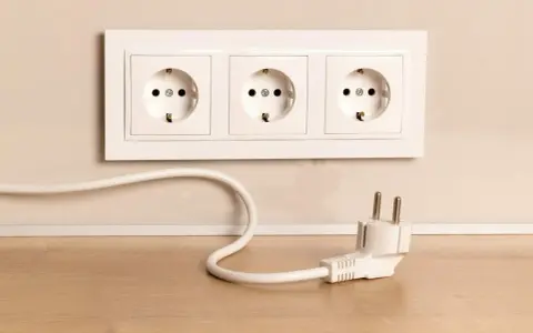 Biały kabel z wtyczką leży na drewnianym blacie obok potrójnego gniazdka. Jak schować kable, by wyglądały estetycznie?