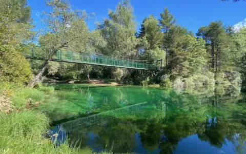Puente verde cruza un río de aguas cristalinas y verdes, reflejando el bosque exuberante. Un lugar idílico para explorar el **alto tajo**.