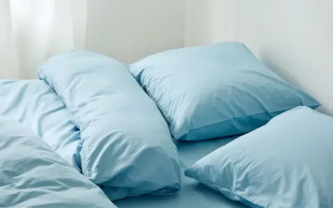 En bädd med ljusblå sängkläder, troligen percale eller satin, med flera kuddar och ett täcke.