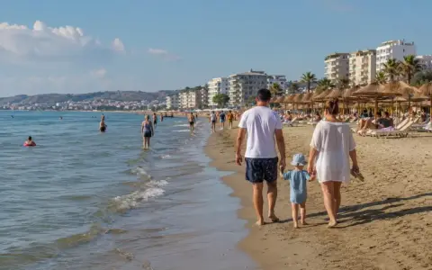 Rodzina spaceruje po piaszczystej plaży w Durrës. W tle widać morze, budynki i parasole plażowe.