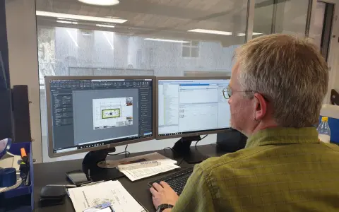 Mann am Computer, der Baupläne nach DIN 4426 erstellt. Auf dem Bildschirm sind CAD-Zeichnungen und eine Liste von Bauteilen zu sehen.