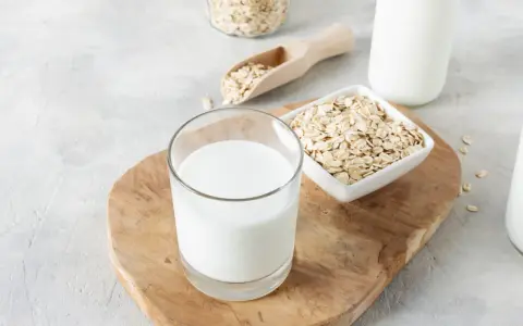 Ein Glas cremiger Hafermilch steht auf einem Holzbrett, daneben Schüsseln mit Haferflocken. Hafermilch gesund und lecker!