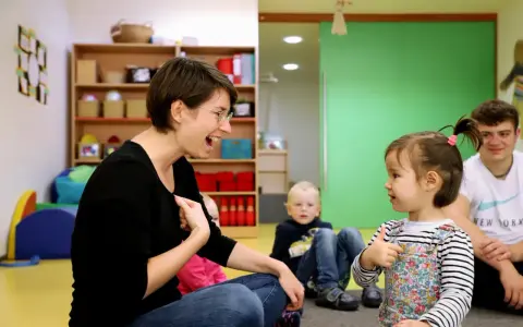 Eine Frau lacht und gestikuliert, während sie mit einem Kleinkind sprechen übt. Ein Junge und ein Mann sitzen im Hintergrund.