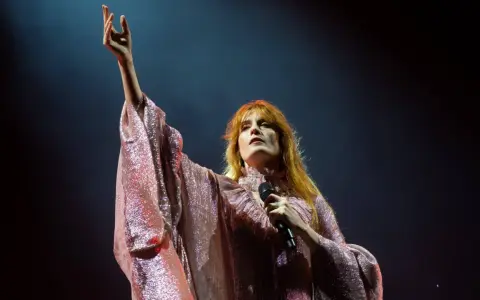 Florence Welch z Florence and the Machine na koncercie, w błyszczącej sukni, z uniesioną ręką.