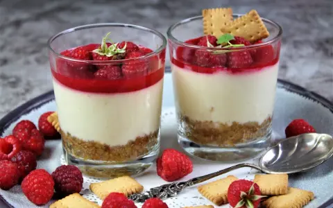 Zwei Gläser mit kalorienarmen Desserts im Glas: cremige Schicht, Himbeersoße und Keksboden. Garniert mit frischen Himbeeren und Keksen.