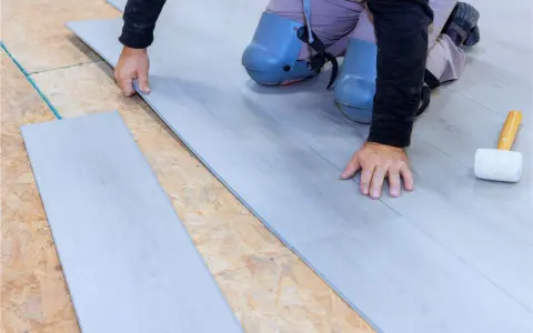 Man con rodilleras coloca cuidadosamente tablones de suelo laminado gris sobre una base de madera.