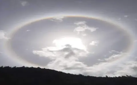 Un alone arcobaleno circonda il sole tra le nuvole, creando un effetto magico sopra una collina scura.