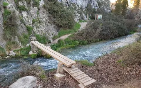 Puente de madera sobre un río caudaloso, parte de la ruta del zarzalar. Senderos y vegetación agreste flanquean el camino.