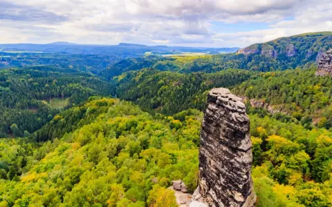 Skalny filar góruje nad zielonymi lasami w Czechach, blisko granicy. Krajobraz pełen jest drzew i skalistych zboczy.