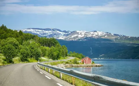 En slingrande väg längs en fjord i Norge, med snöklädda berg i bakgrunden. En röd stuga vid vattnet. Perfekt för en roadtrip norge rutt.