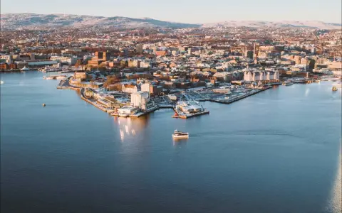 Widok na zimowe Oslo z wody. Mała łódka płynie po spokojnym akwenie, w tle rozciąga się miasto i ośnieżone wzgórza.
