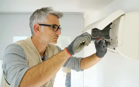 Tecnico con occhiali e guanti sta per smontare split condizionatore dal muro con un cacciavite.