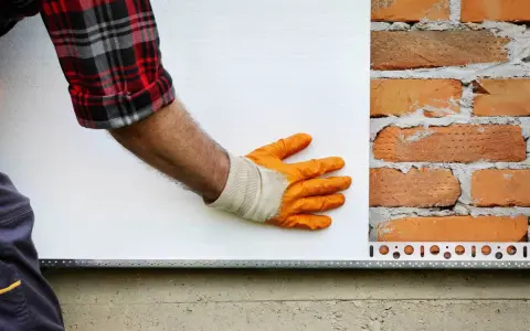 Ręka w pomarańczowej rękawicy przykłada płytę pir do ceglanej ściany.