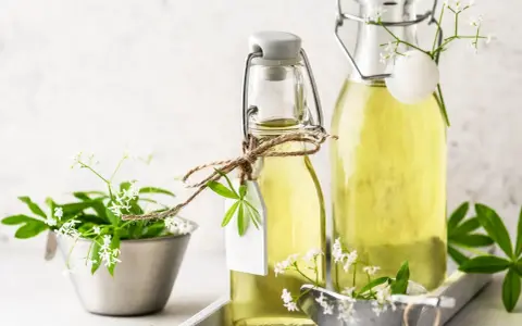 Zwei Flaschen mit selbstgemachtem Waldmeistersirup, dekoriert mit frischen Waldmeisterzweigen und kleinen weißen Blüten.