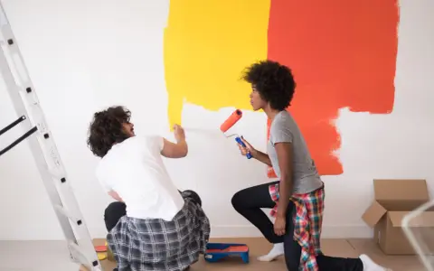 Un couple peint un mur en jaune et orange. Ils utilisent des rouleaux pour nettoyer peinture glycero. Une échelle est à gauche.